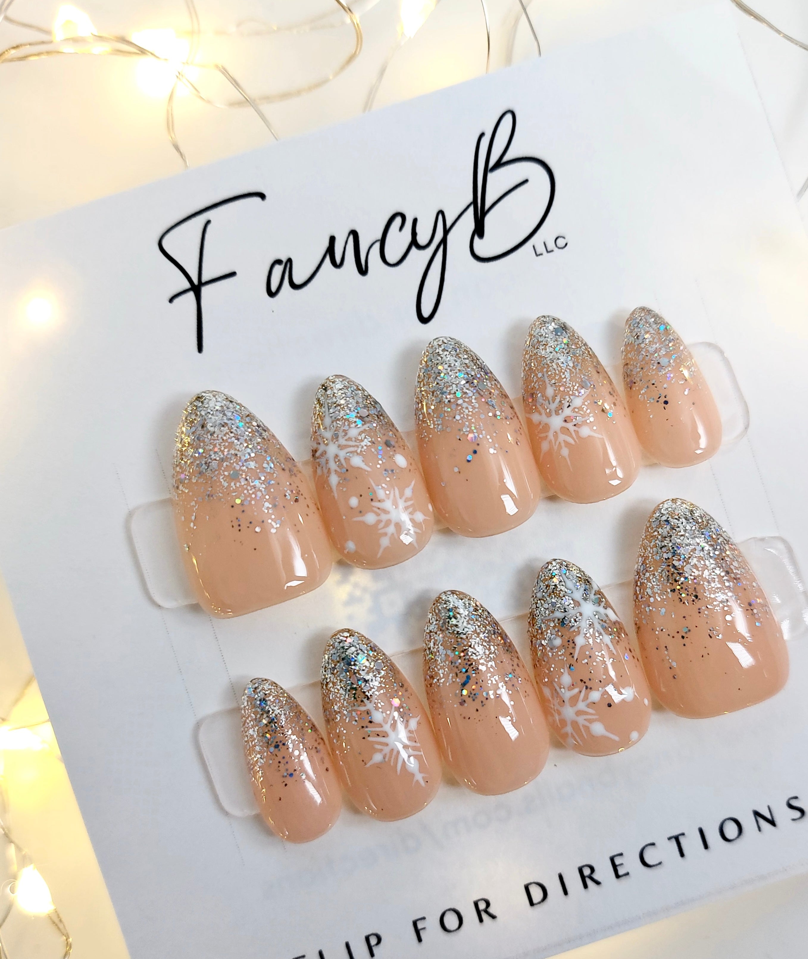 Custom press on nails with nude color, silver glitter ombre tips and hand painted snowflake designs on a short almond shape. FancyB Nails.