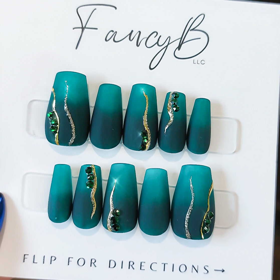 teal tropical nails with ombre, gold chrome lines, gold glitter, and emerald gems in a matte finish.