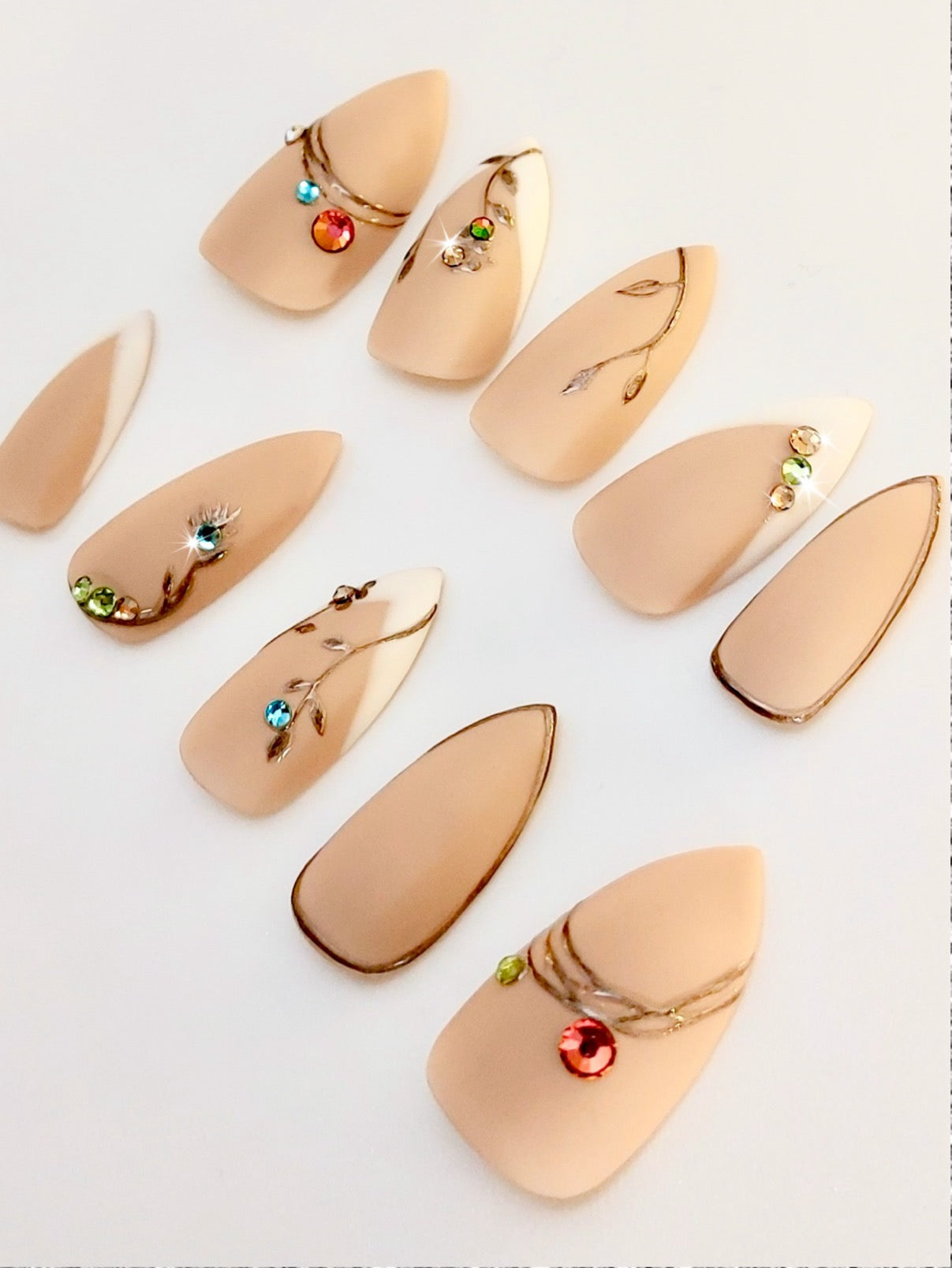 Custom press on nails with nude matte, gold metallic accents, colorful gems like christmas ornaments and french tips on short sharp stiletto shape. FancyB Nails.