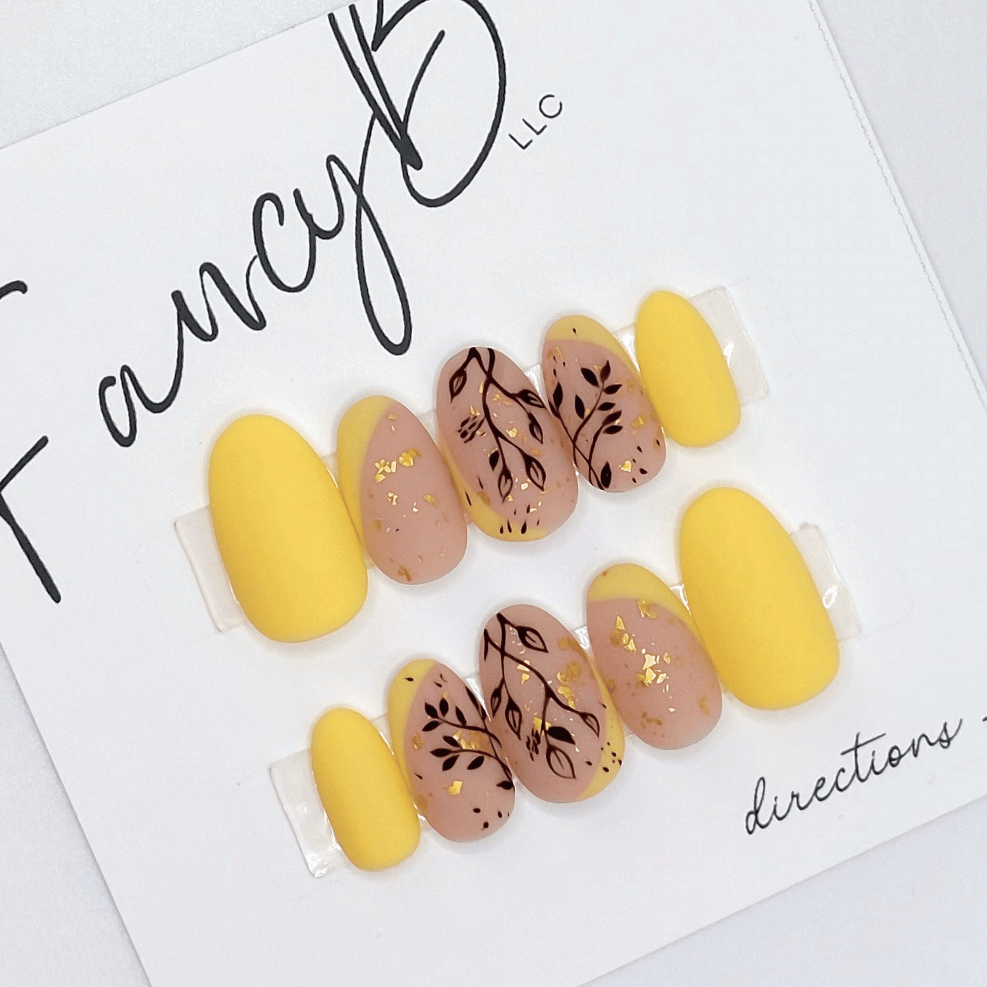 Custom press on nails with light yellow french tips, black floral designs, gold flakes, and a nude base with matte finish and short oval shape. Custom nails from FancyB.