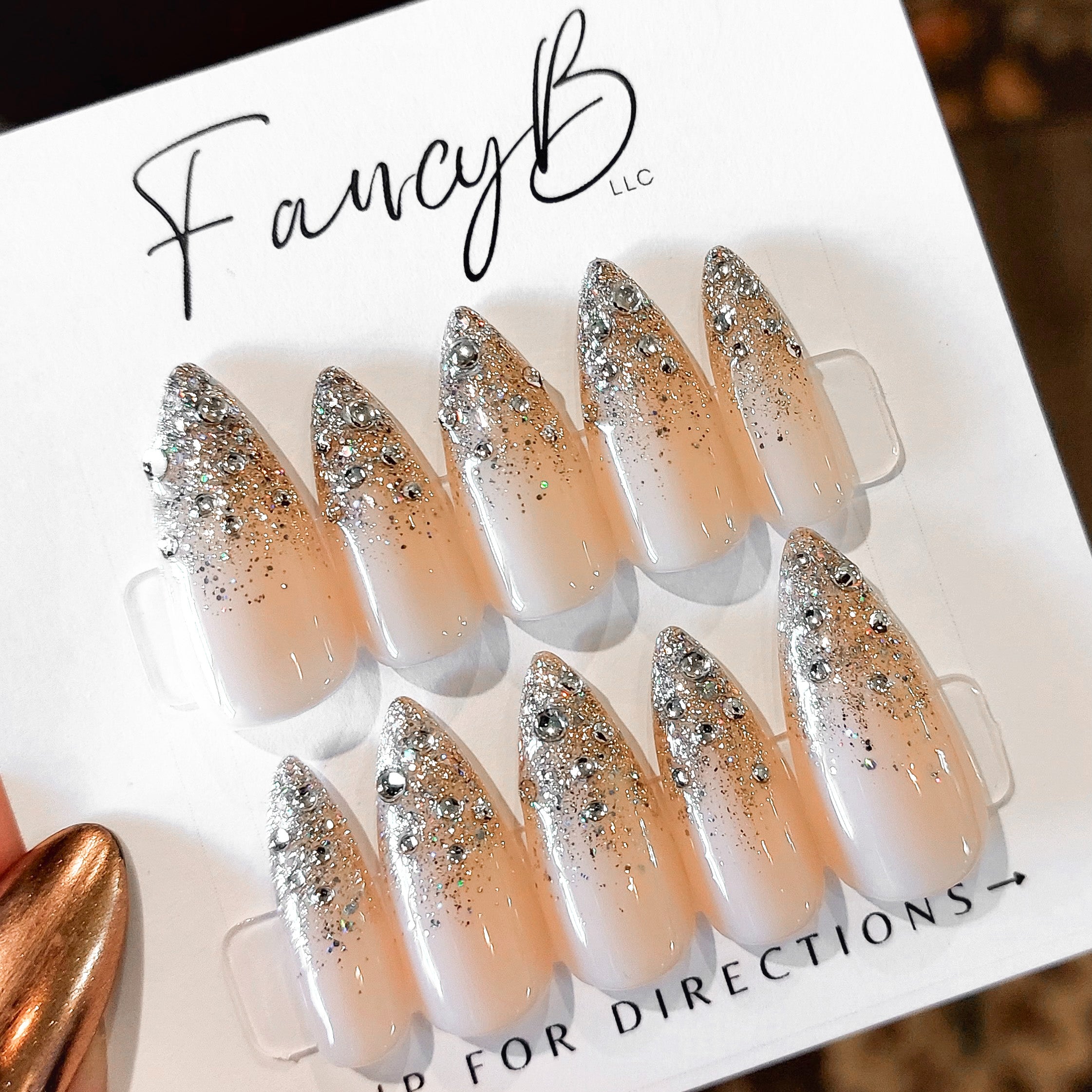 Custom press on nails with nude base color, silver gems and silve glitter ombre on sharp stiletto nail shape. FancyB Nails.