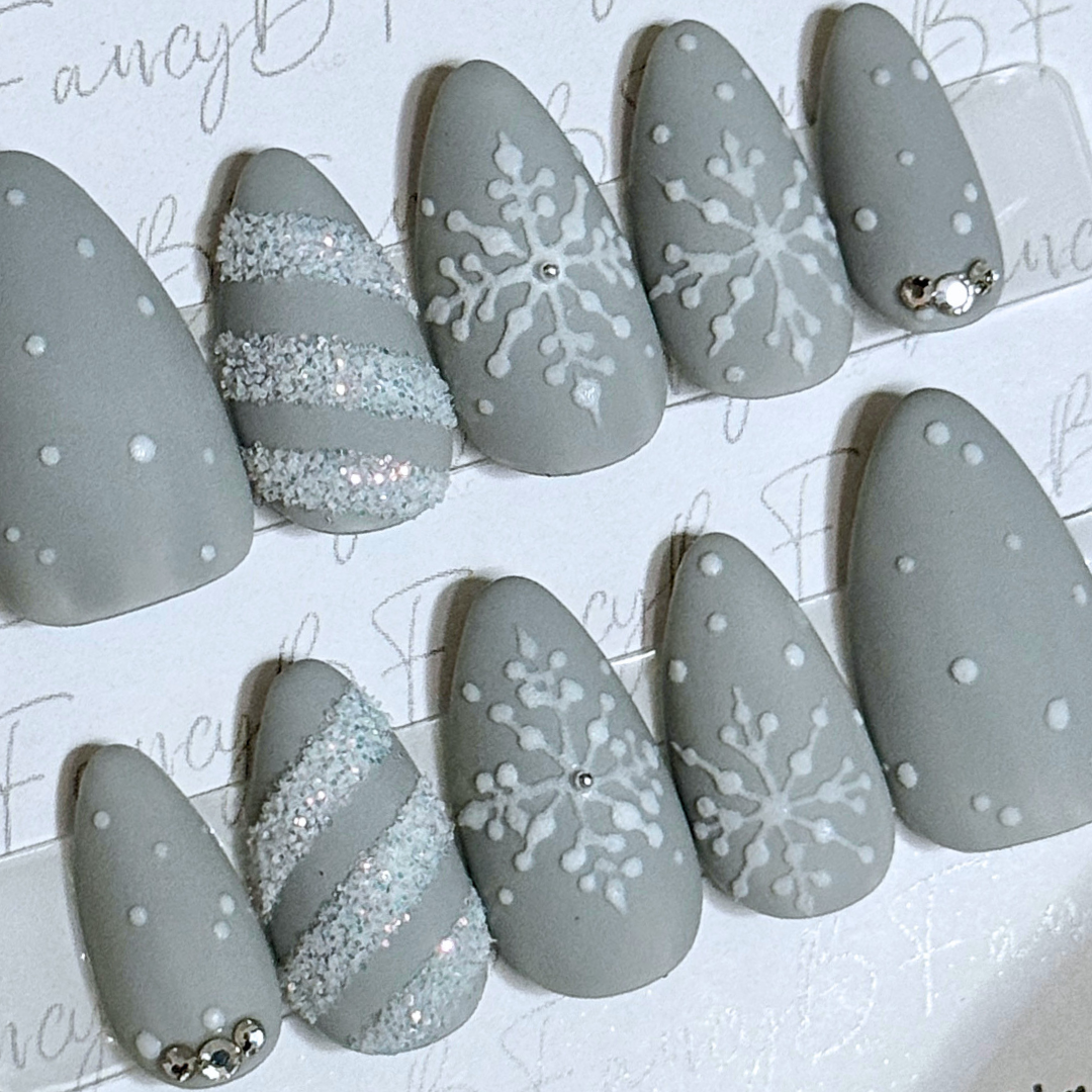 snowflake press on nails with white hand painted snowflakes, white glitter candy cane stripes, light grey background color, and diamond gems on short almond nails by FancyB Press on Nails.