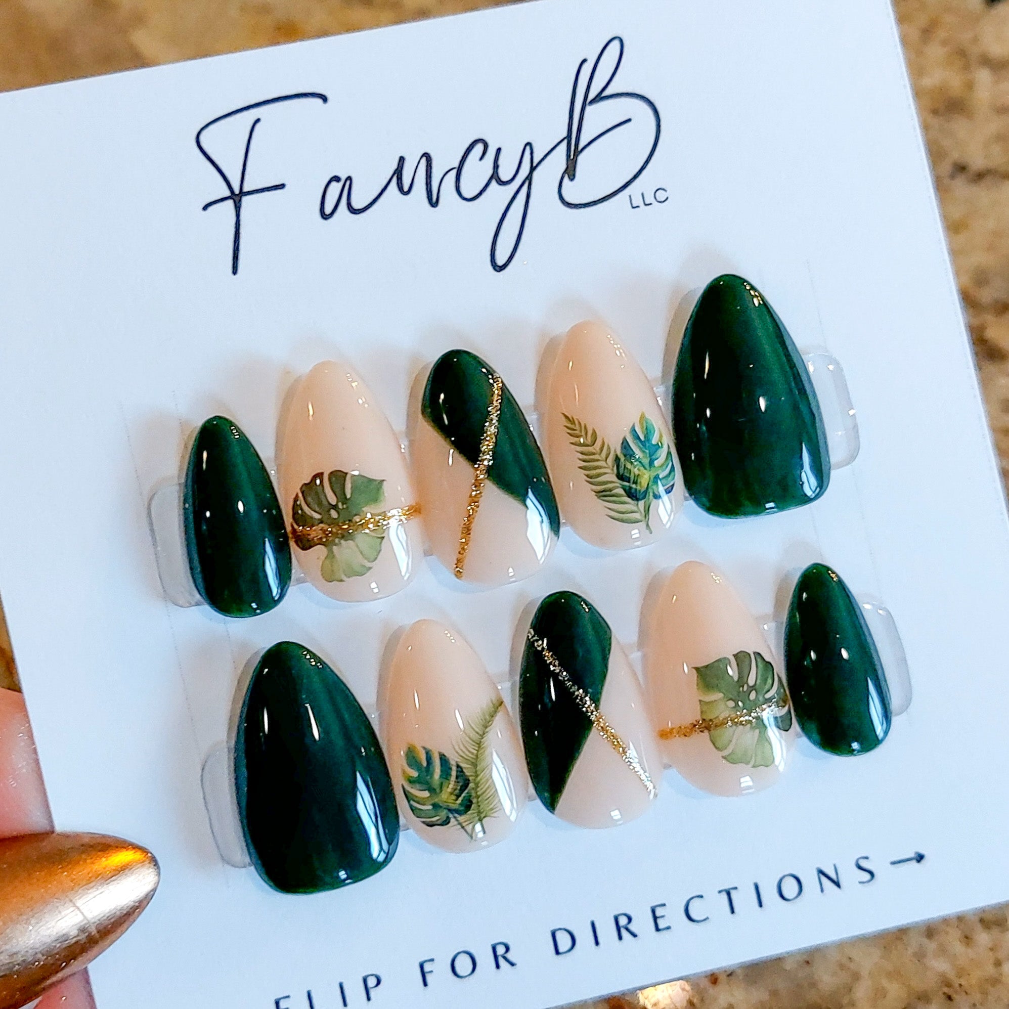 Custom press on nails with emerald green accents, monstera and palm leaves, gold glitter lines and a nude base color on a short almond shape. FancyB Nails.