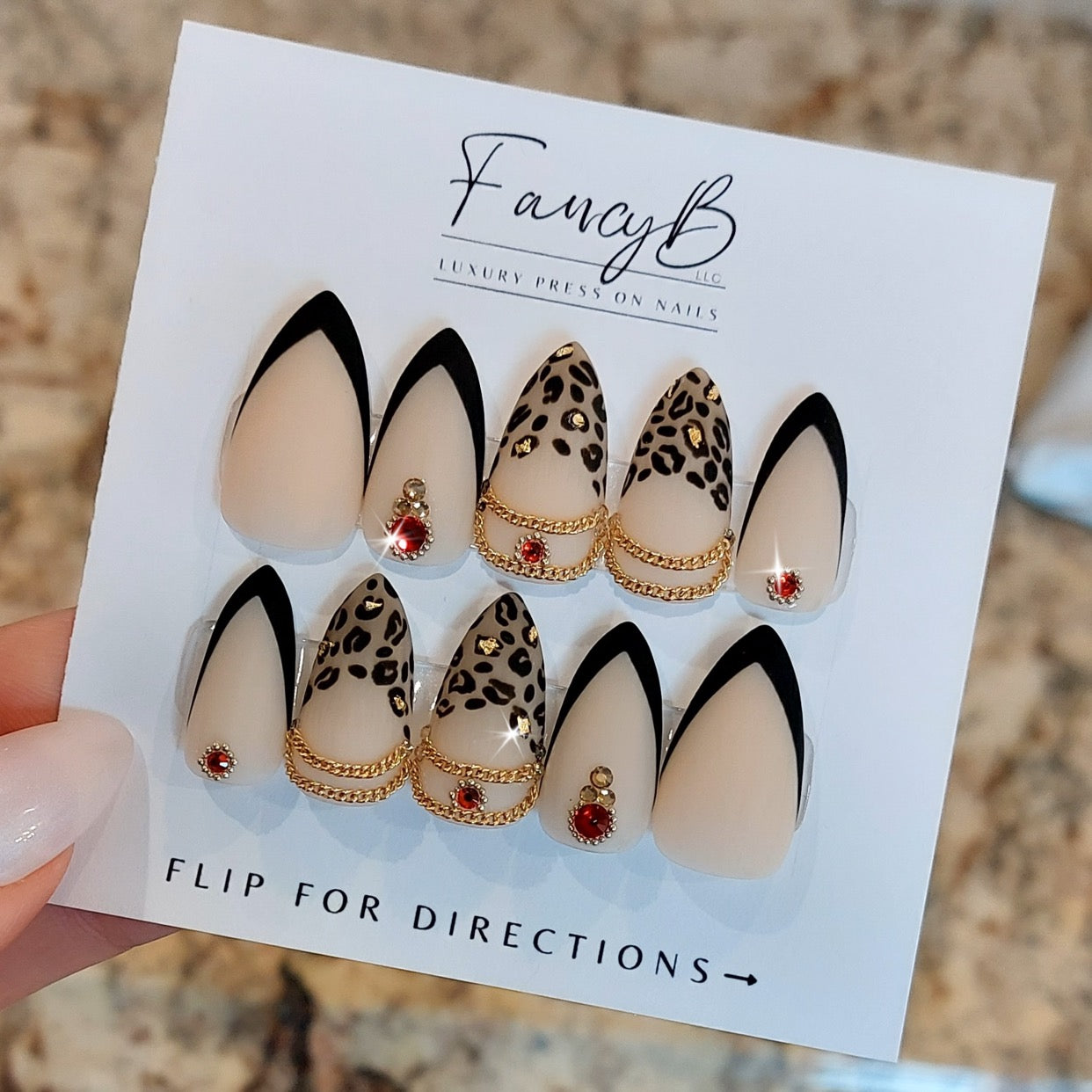 custom press on nails in sharp stiletto shape with leopard tips, gold flakes, black and neutral v-french, ruby red gems, and gold chains.