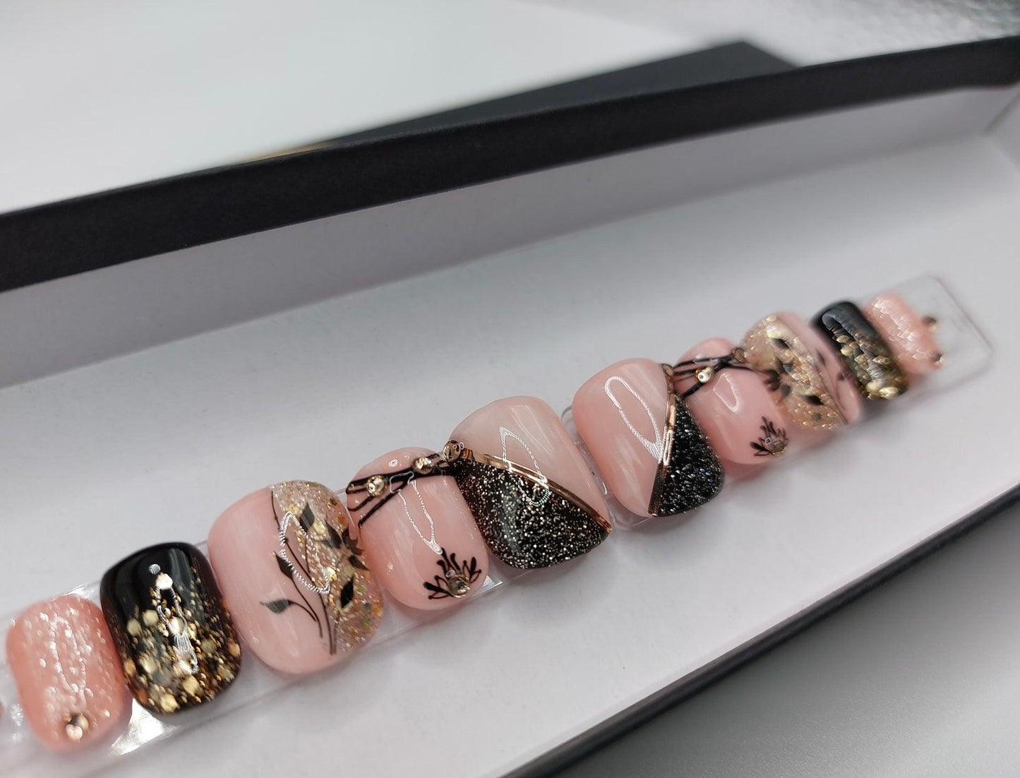 Custom nail design in a soft rose quartz base color with black and gold glitter accents, floral designs, champagne gems and a short squoval shape.